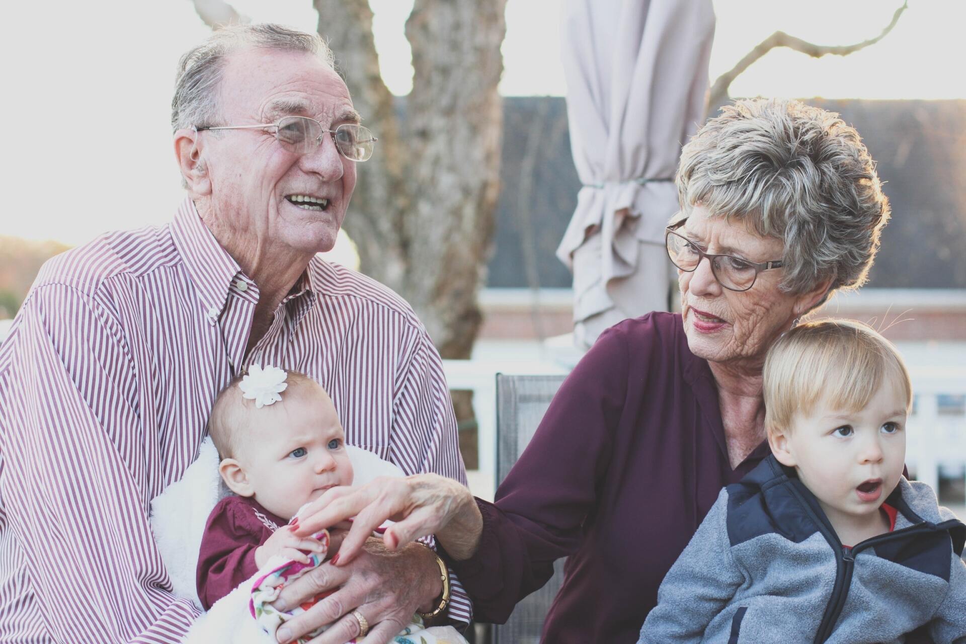 An elderly man and woman, part of the Sandwich Generation, sit outdoors holding a baby and a toddler. Smiling warmly, they exemplify caregiving as they interact with the children and navigate life’s precious moments together.