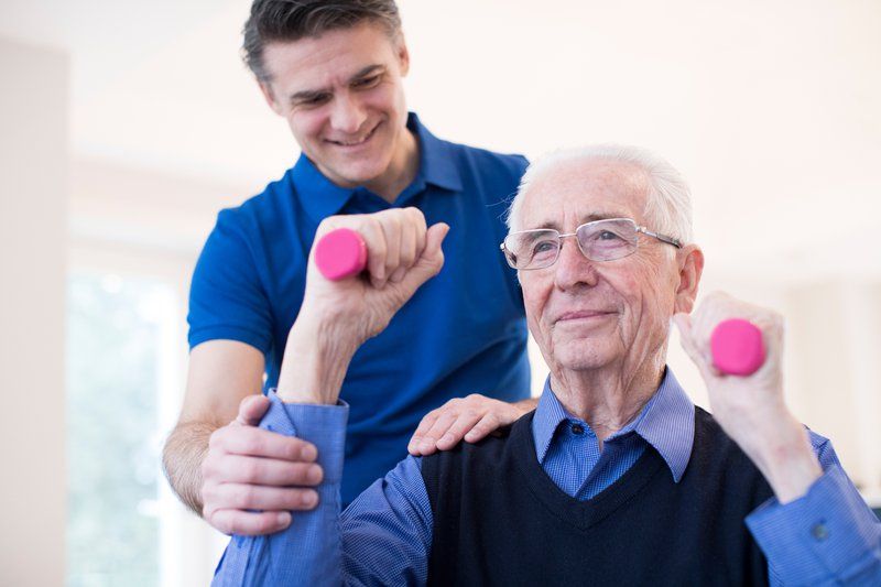 An older man lifts small pink dumbbells as part of his exercise routine, while a younger man in a blue shirt assists and supports him for effective dementia management.