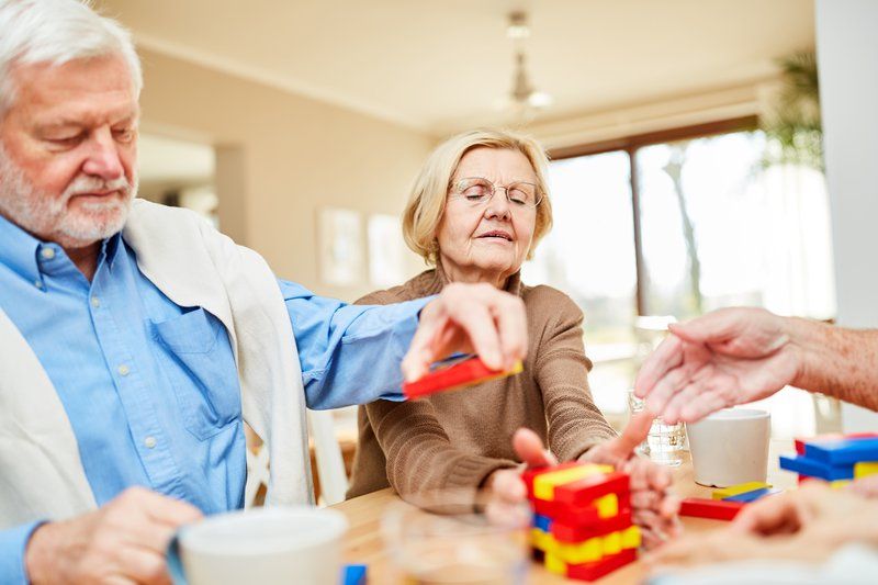 Three older adults sit at a table playing a game with colorful stacking blocks, enhancing their social skills as they interact. Cups and hands are visible on the table.