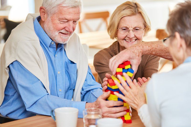 Three older adults sit at a table, smiling and playing a game with colorful wooden blocks—a glimpse into innovative dementia care that encourages connection and joy. Cups and glasses rest nearby as part of their supportive care plans.
