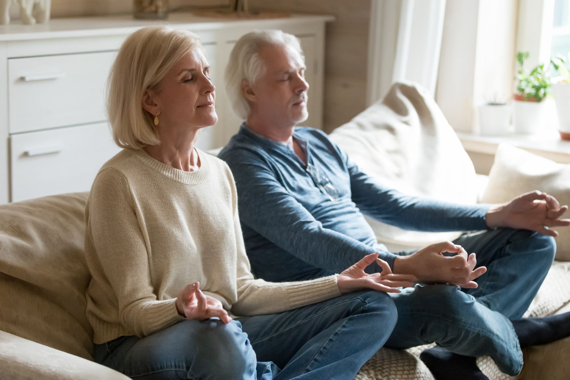 Two older adults sit cross-legged on a couch indoors, meditating with eyes closed and hands on their knees—embracing mindfulness practices known for their benefits, especially among dementia patients.