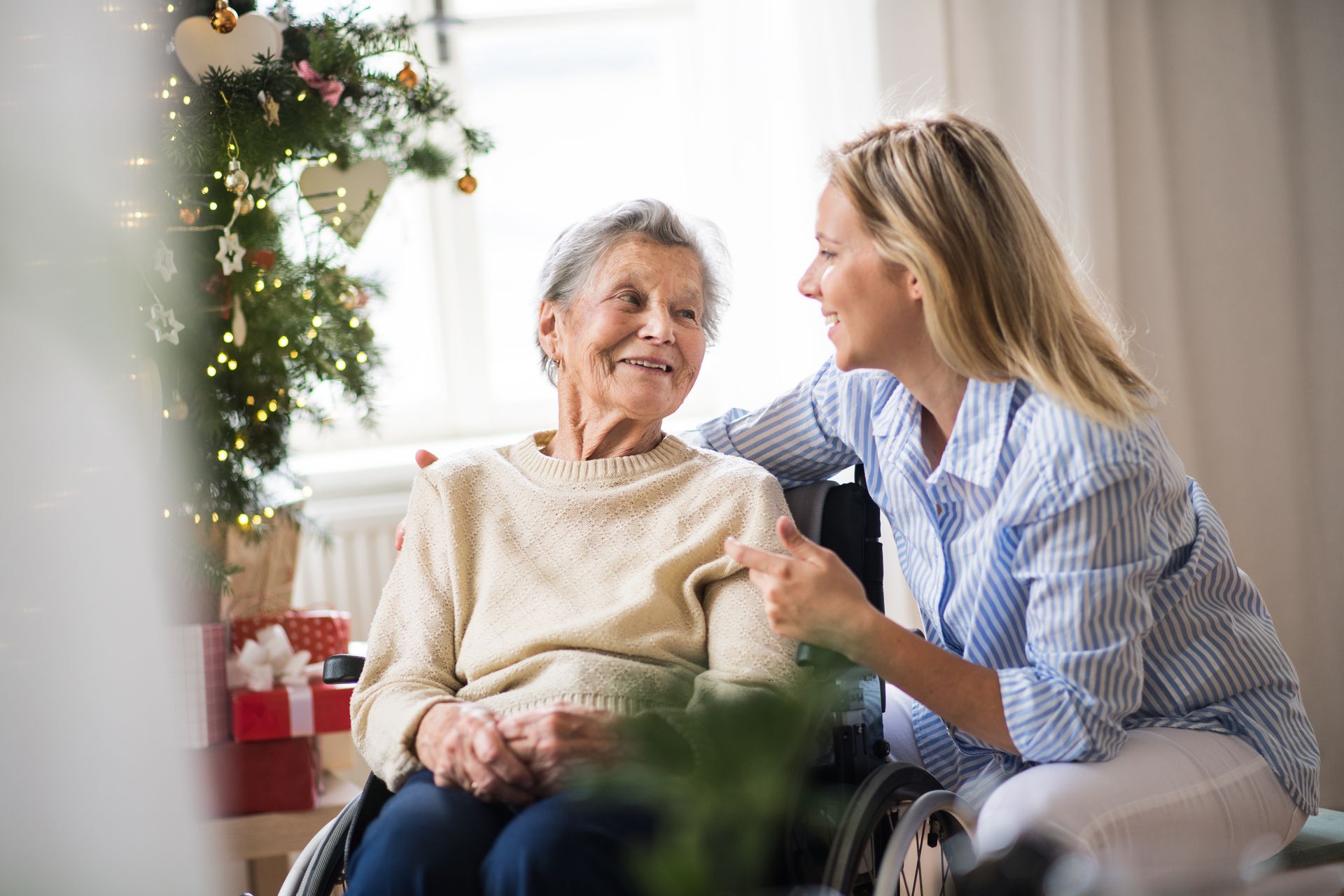 An older woman in a wheelchair and a younger woman smile and talk beside a decorated Christmas tree indoors, highlighting the warmth of social connections for seniors during the winter months.