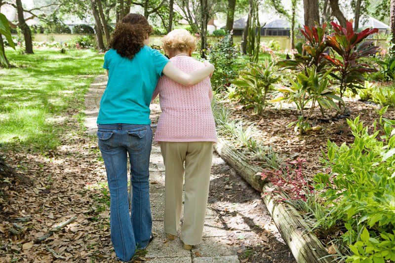 Two women, one younger and one older, walk arm-in-arm along a garden path surrounded by greenery and trees on a sunny day, reflecting the best care for cognitive impairment through companionship and support.