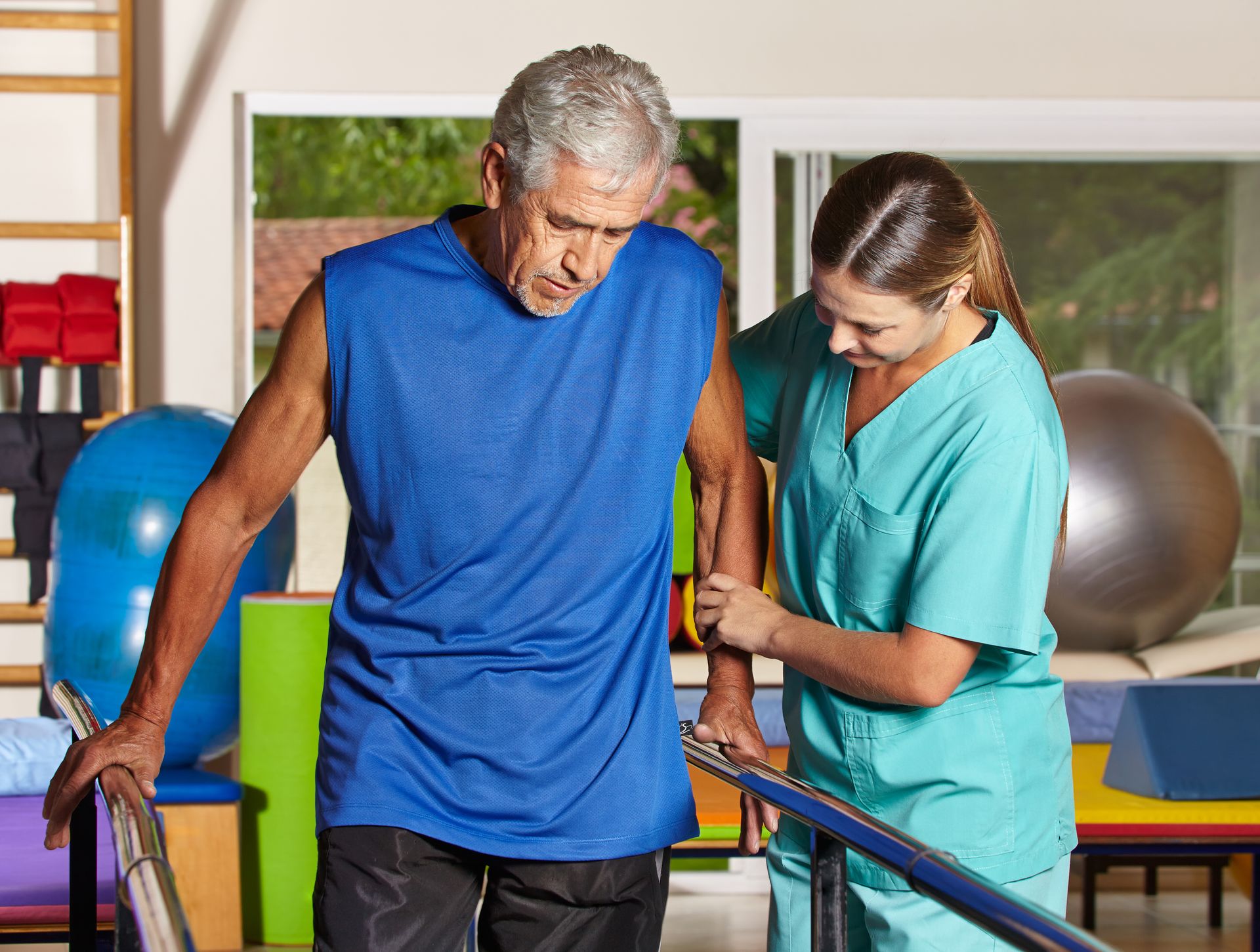 An older man practices walking between parallel bars with assistance from a healthcare professional and adaptive equipment in a physical therapy setting.