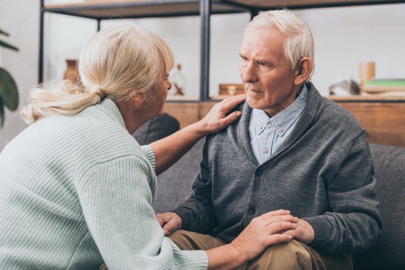 An elderly woman sits beside an elderly man on a couch, gently touching his shoulders and knees while speaking to him—offering Alzheimer's support as he faces the challenges of the disease, his expression serious and distant.