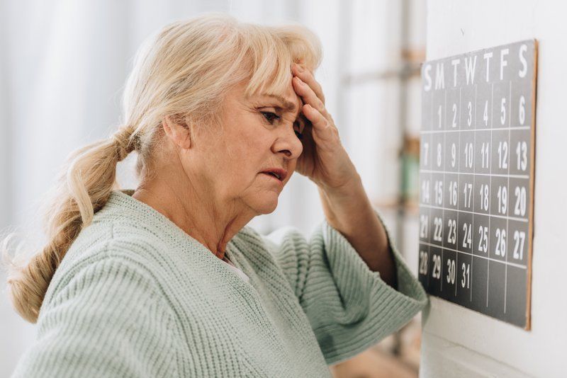 An elderly woman with a concerned expression stands in front of a wall calendar, resting her hand on her forehead—she may be experiencing early signs of cognitive impairment.