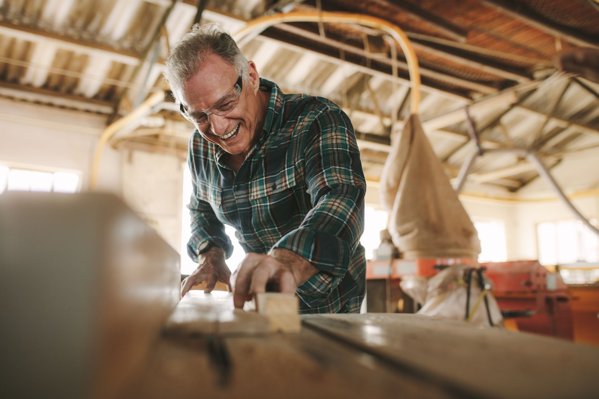 An older man wearing glasses and a plaid shirt works with wood in a workshop, smiling as he sands a piece of lumber, preparing for returning to work after brain injury.