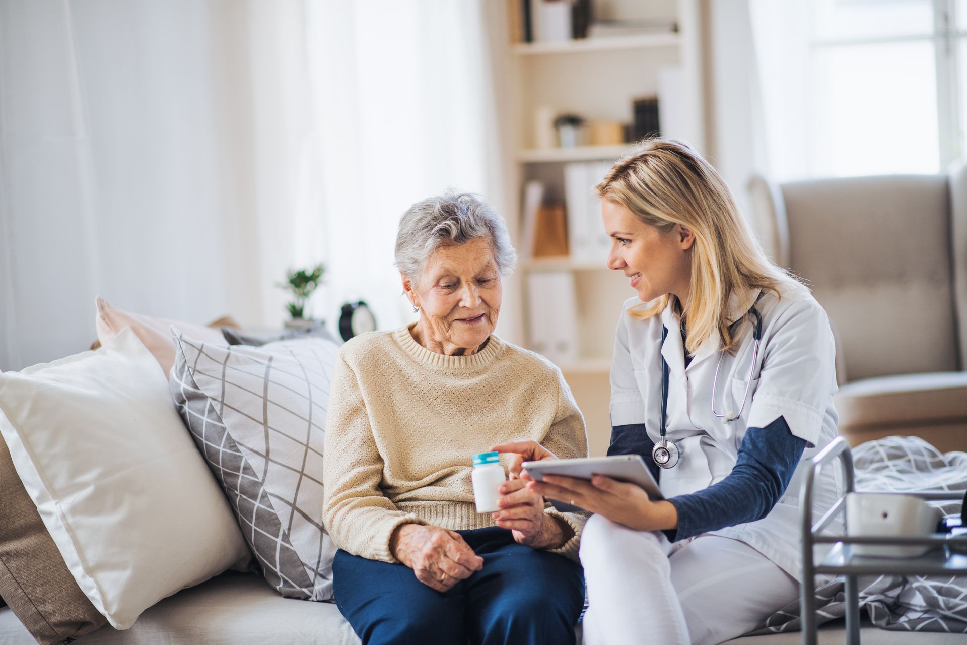 A nurse in uniform shows a tablet to an elderly woman holding a medication bottle, supporting her in navigating brain injury while they sit together on a couch in a bright living room.
