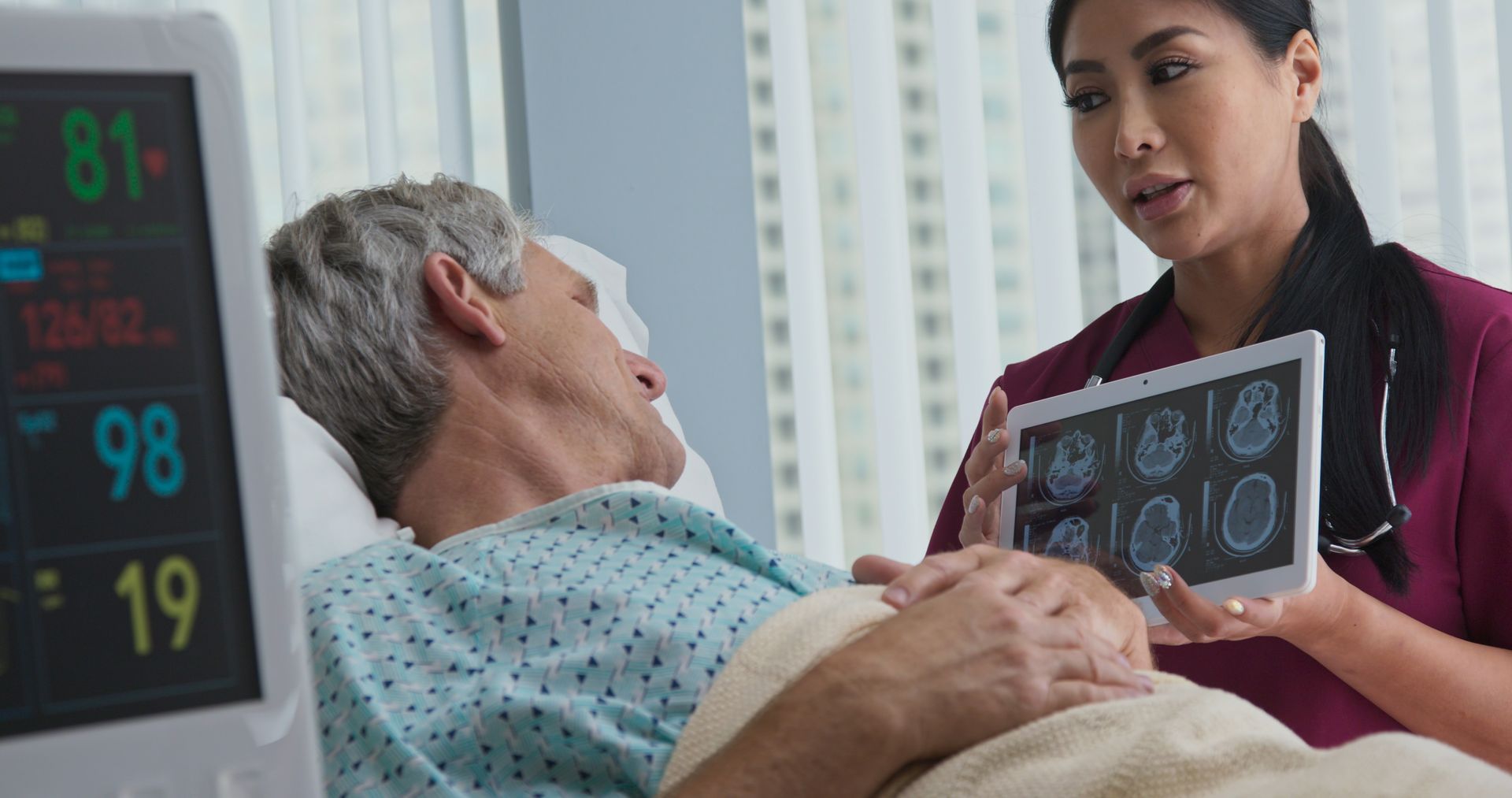 A healthcare professional shows a tablet with brain scan images to an older patient with a traumatic brain injury lying in a hospital bed, with monitoring equipment visible.