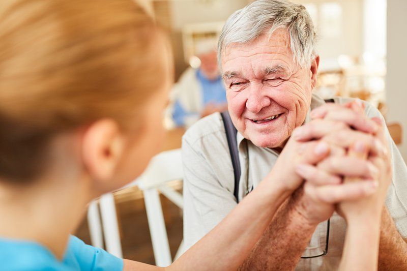 An older man smiles while holding hands with a caregiver in a bright indoor setting, reflecting warm support for those living with dementia.