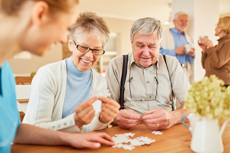 Two seniors and a caregiver assemble a puzzle at a table, enjoying Alzheimer's support, while another older couple converse and drink coffee in the background.
