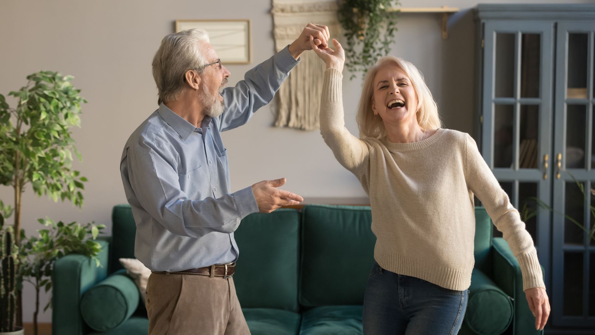 An older man and woman are smiling and dancing together in a cozy assisted living space, surrounded by green plants, a green sofa, and a blue cabinet in the background.