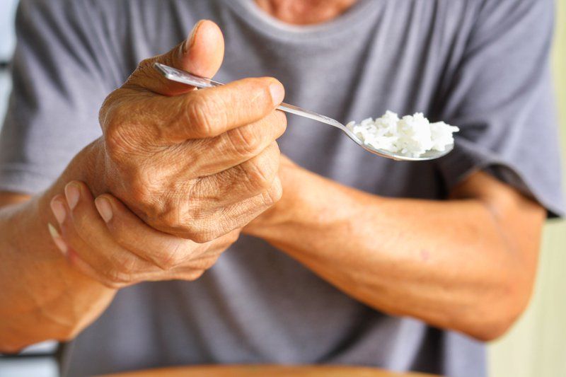 An older person holds their wrist with one hand while eating white rice with a spoon, suggesting difficulty or discomfort in movement—highlighting the importance of Parkinson’s support and personalized care in daily activities.