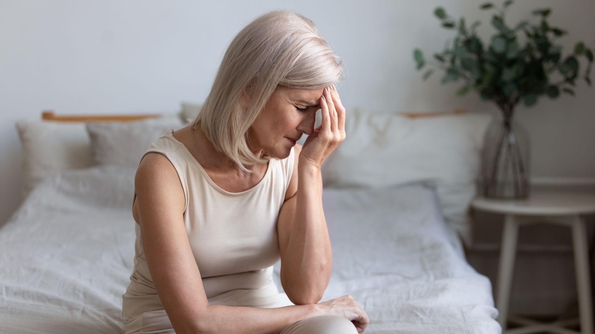 An older woman sits on a bed with her hand on her forehead, appearing distressed or deep in thought—possibly struggling with chronic pain after brain injury—with a plant and side table in the background.