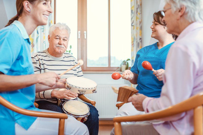 Four people, including two older adults and two caregivers, sit in a circle playing percussion instruments in a brightly lit room, sharing music as a creative activity for dementia and discovering moments of joy in dementia together.