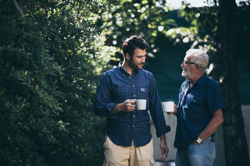 Two seniors stand outdoors holding white mugs, engaged in conversation, sharing their stories amid greenery and sunlight in the background.