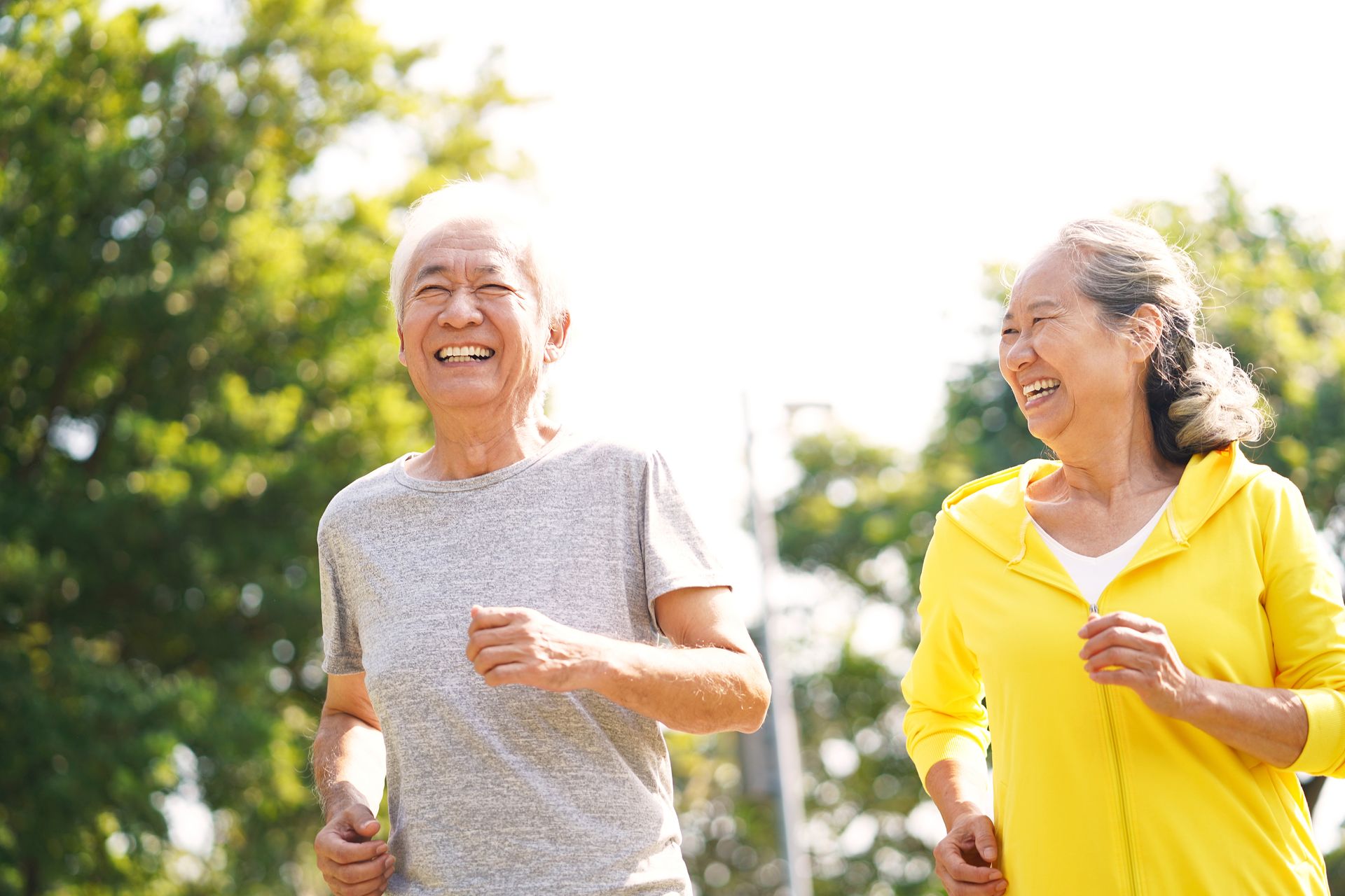 Two older adults are jogging outdoors on a sunny day, smiling and wearing casual athletic clothing with trees in the background, enjoying the connection and brain health benefits of physical activity.