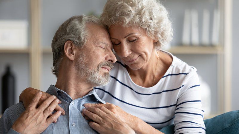 Elderly couple sitting close together with eyes closed, gently embracing and touching foreheads in a moment of compassion and affection.