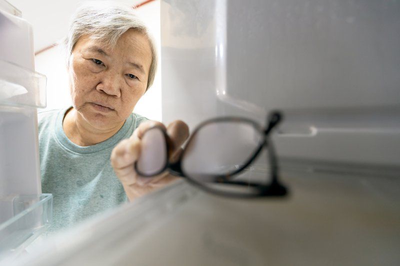An older person retrieves a pair of eyeglasses from inside a refrigerator, highlighting some behavioral challenges often encountered when managing Alzheimer’s care, viewed from the perspective of the fridge interior.
