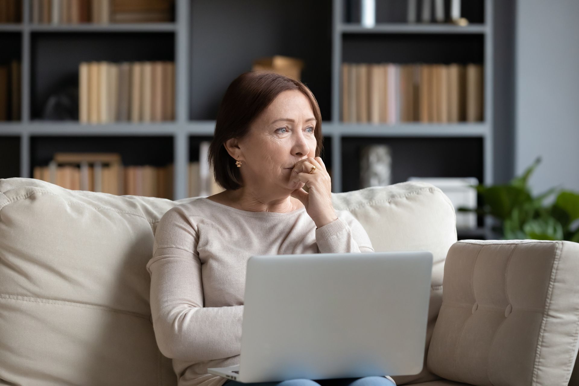Middle-aged woman sitting on a couch with a laptop, looking thoughtful as she explores ways to support senior grief in a cozy living room with bookshelves in the background.