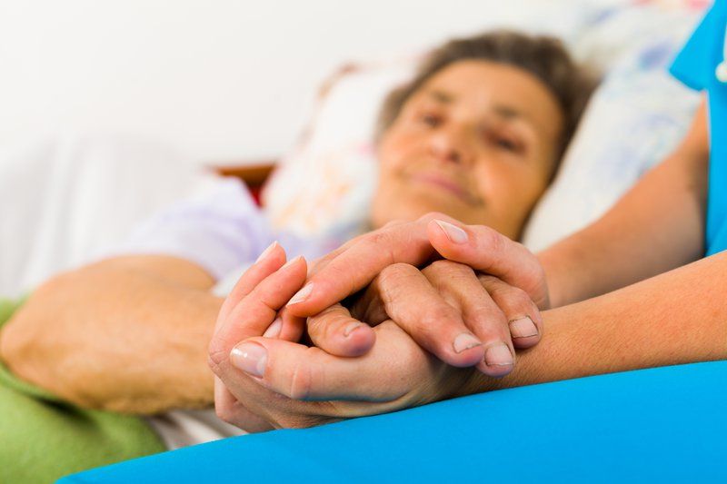 An elderly person with Parkinson's lies in bed, holding hands with a caregiver who offers comfort and support, embodying holistic care for seniors.
