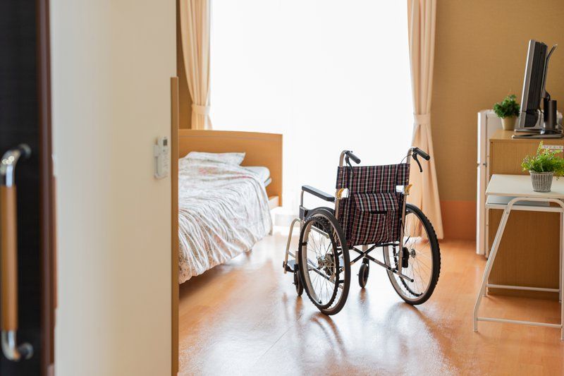 A wheelchair is positioned near a bed in a bright, neatly arranged bedroom with light curtains and wooden furniture, reflecting the comfort and support found in quality assisted living settings.
