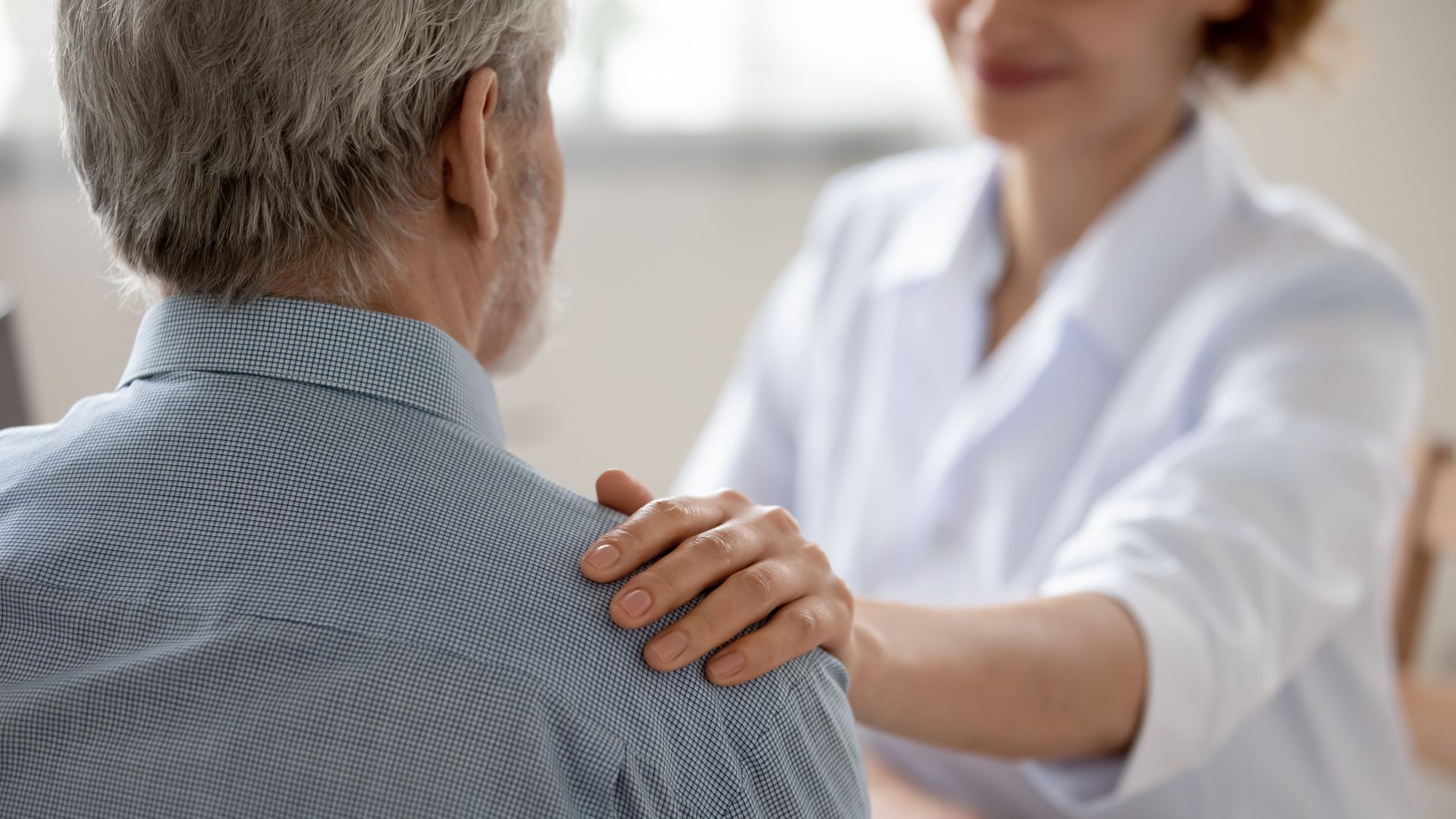 A healthcare professional rests a reassuring hand on an older man's shoulder during a conversation, offering social support in a bright, indoor setting.