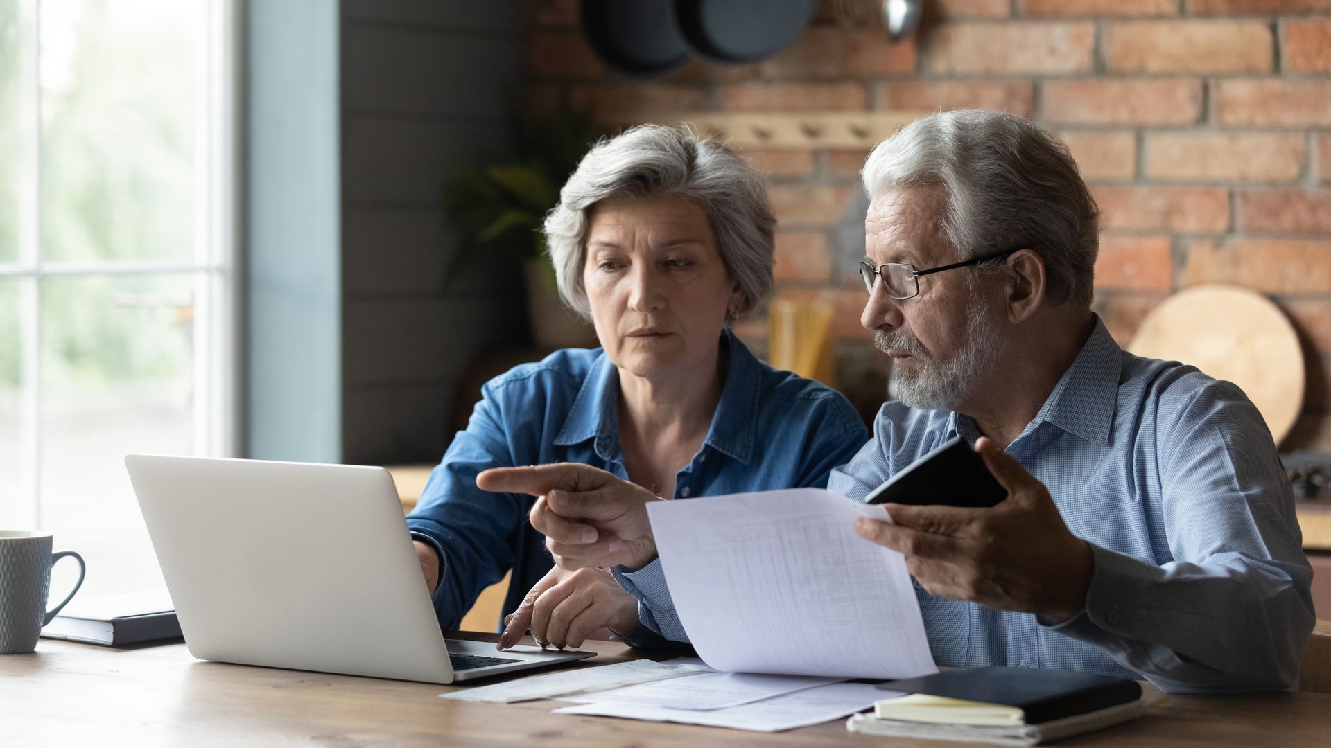 Two older adults sit at a table with a laptop, documents, and a phone, appearing focused as they review paperwork together for tax season and discuss financial planning.