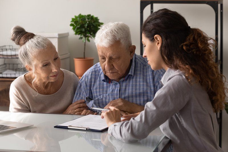 An elderly couple sits at a desk with a young woman, reviewing financial planning documents together; the man holds a pen, ready to sign a form.