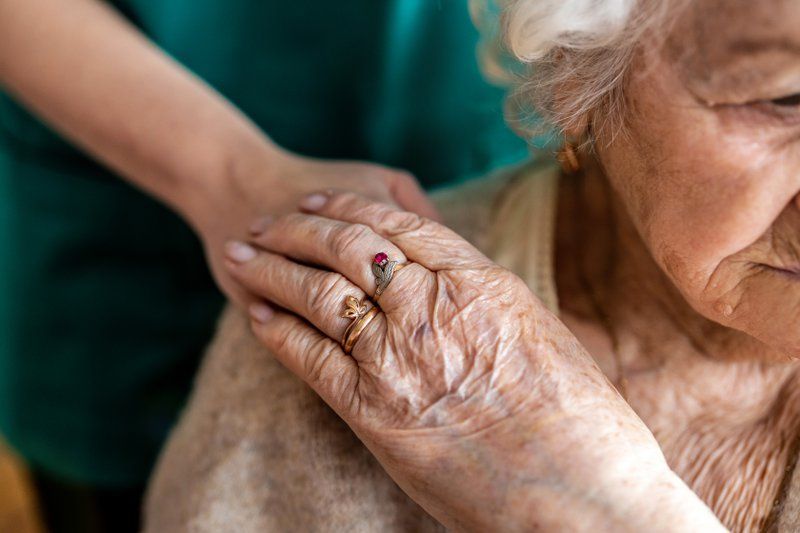 A younger person rests a hand on the shoulder of an elderly woman wearing rings, offering comfort or support and enhancing her quality of life during Frontotemporal Dementia (FTD) care.