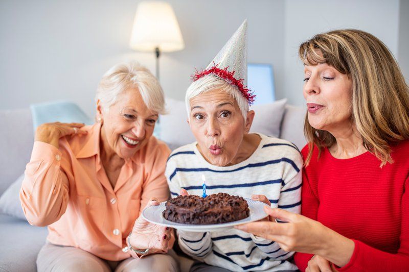 Three women sit together on a couch at Assured Senior Living, one wearing a party hat and blowing out a candle on a chocolate cake while the others smile and watch during this joyful celebration of life.