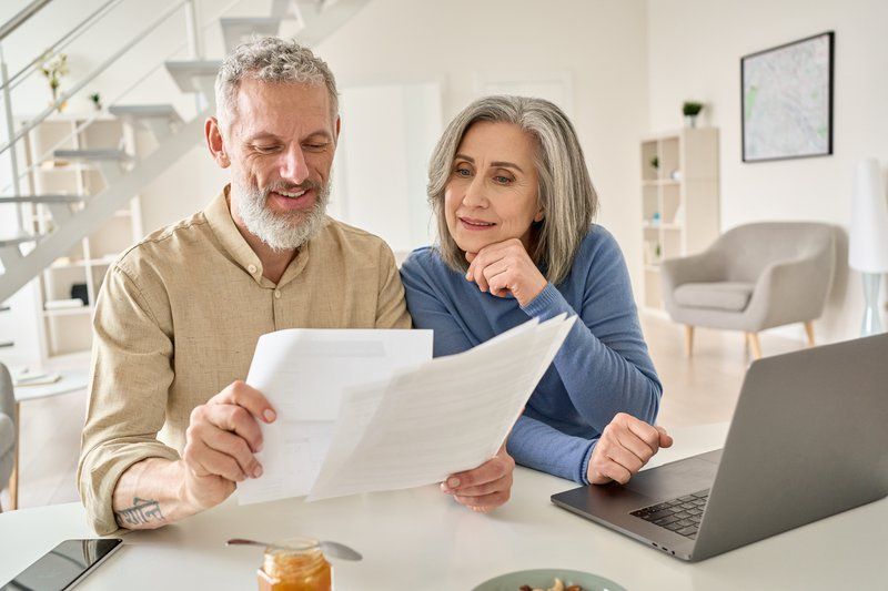 Older couple sitting at a table, looking at documents together—perhaps discussing legal issues or dementia care—with a laptop open in front of them in a bright, modern living room.