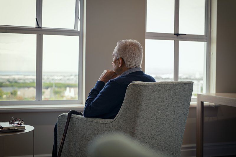 An elderly man with gray hair sits in an armchair by a window, looking outside thoughtfully with a walking cane beside him, reflecting on memories while managing Alzheimer’s and moments of loneliness.