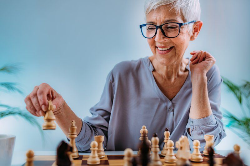 Older woman with short gray hair and glasses smiles while playing chess, holding a white piece above the board—a wonderful way to restore confidence and enjoy social activities after brain injury care.