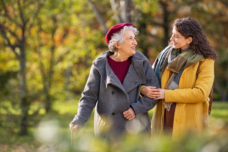 Two women, one older and one younger, walk outdoors in autumn clothing, smiling and holding arms in a park—enjoying the benefits of innovative senior care surrounded by vibrant trees and greenery.