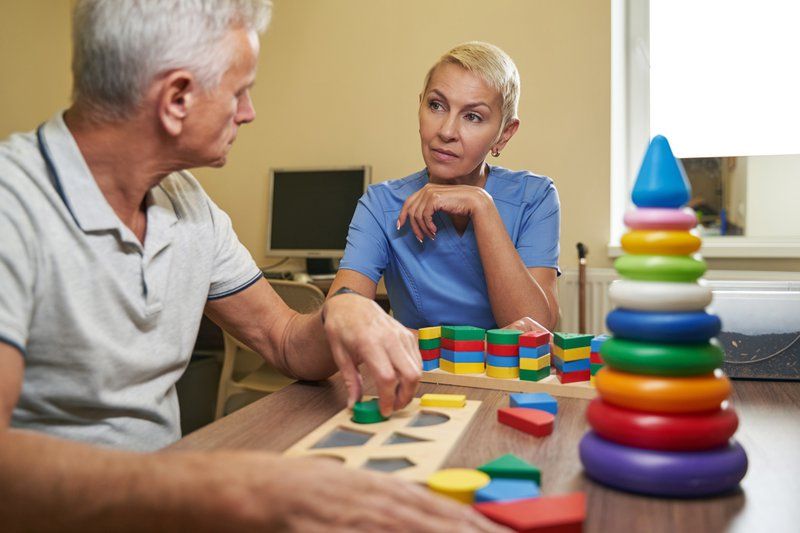 An older man works on shape and color sorting puzzles at a table, with a healthcare professional observing and assisting as part of his personalized care plan.