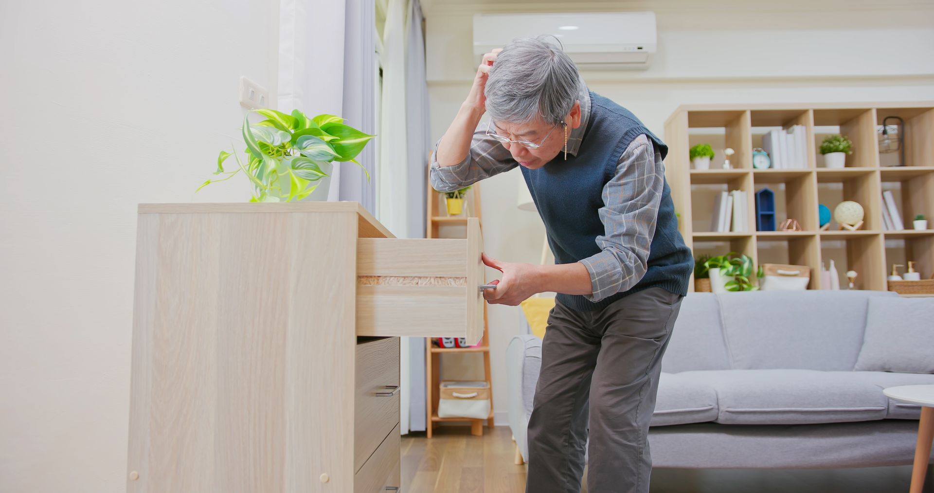 An older adult stands in a living room, looking confused while searching through an open drawer in a wooden cabinet—a scene that may reflect agitation in dementia patients.