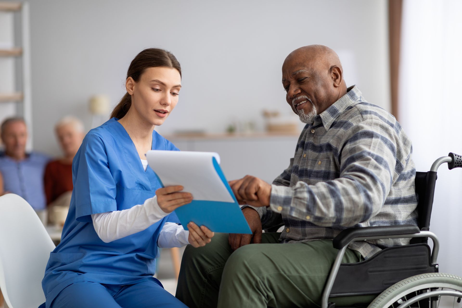 A nurse in blue scrubs shows documents to an older man in a wheelchair, highlighting caregivers supporting Black seniors as they discuss important paperwork in a healthcare setting.