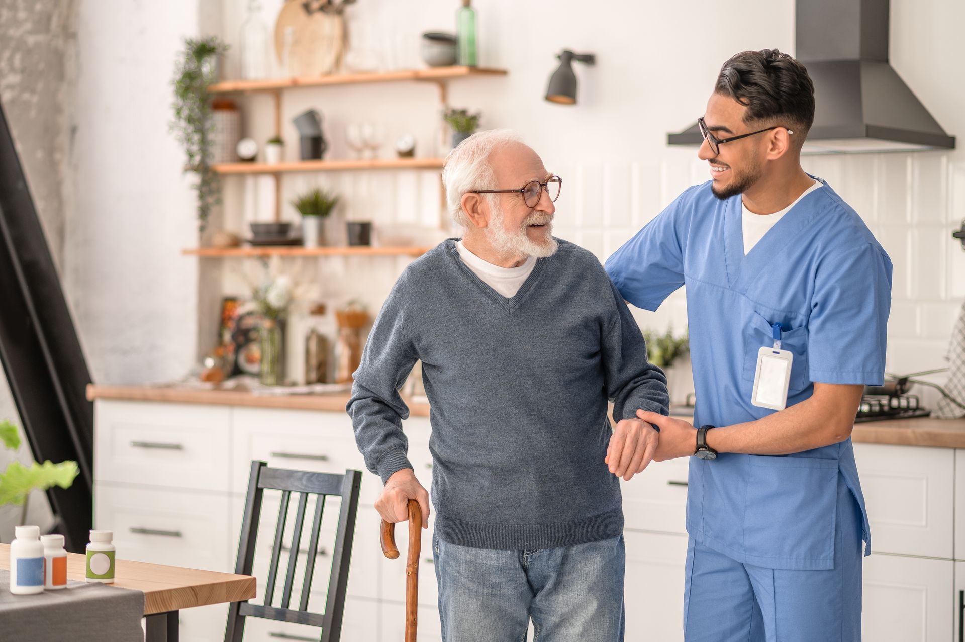 A nurse in blue scrubs assists an elderly man with a cane in a bright, modern kitchen. The man is smiling as the nurse offers supportive arm assistance, demonstrating helpful caregiver tips for patient preparation before medical appointments.