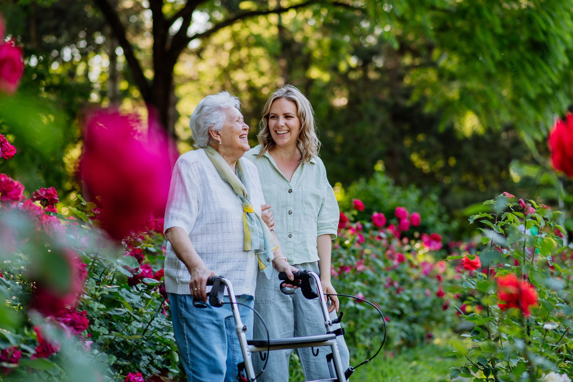 An elderly woman using a walker and a younger woman stand together, smiling in a garden with blooming red roses and lush green trees, illustrating the joy that gardening therapy can bring to brain injury survivors.