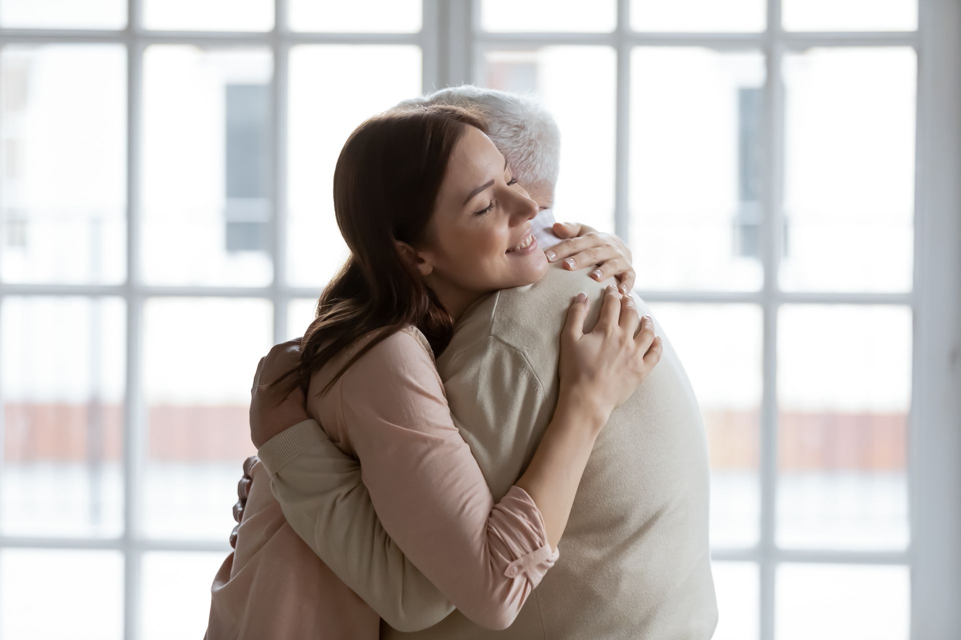 A young woman and an older man, part of the sandwich generation, hug each other warmly in front of large windows, both appearing content and comfortable.