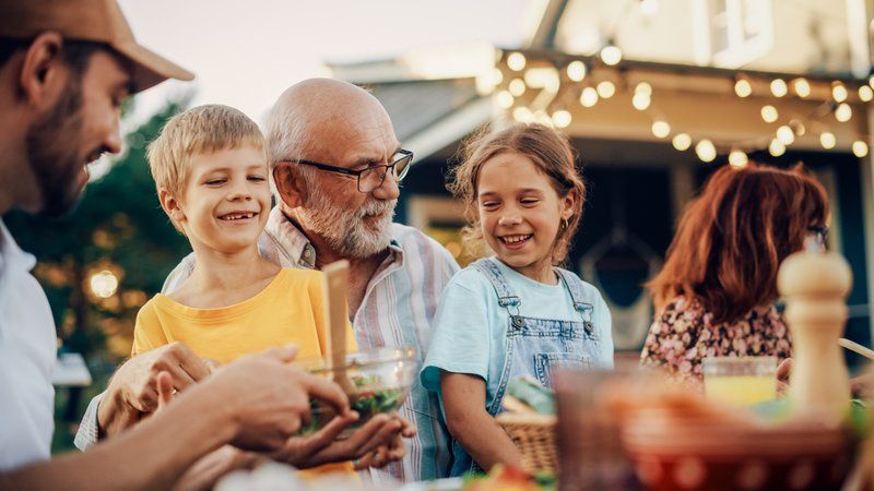 An older man and two children smile together at an outdoor gathering, enjoying food and laughter under string lights—a heartwarming moment that highlights the benefits of intergenerational programs in assisted living communities.