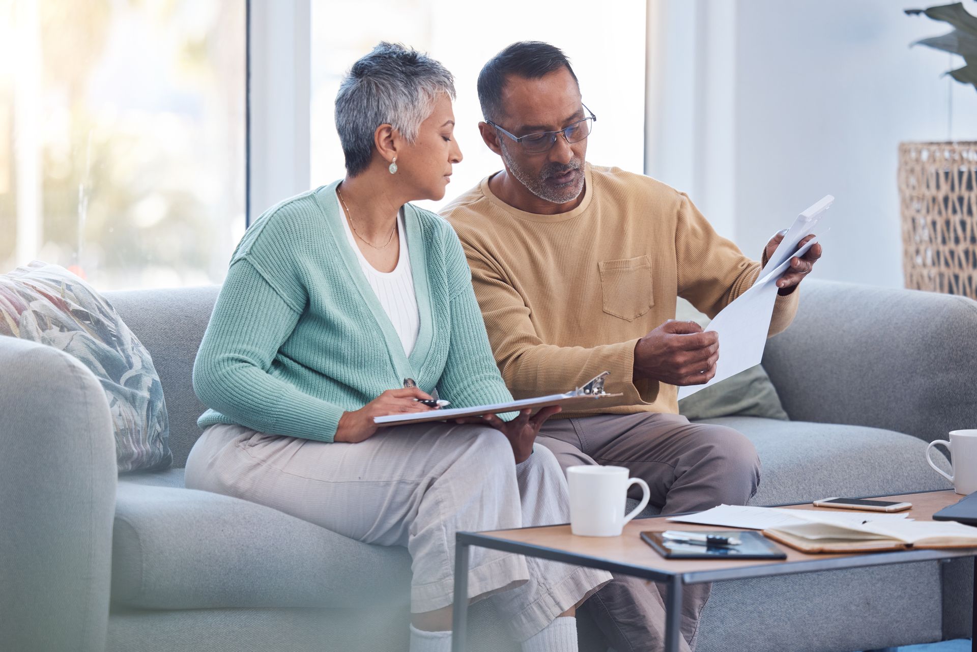 An older couple sits on a couch, reviewing their Elderly Plan documents together. They have a clipboard and papers, with mugs and notebooks from their Business Kit on the table in front of them.