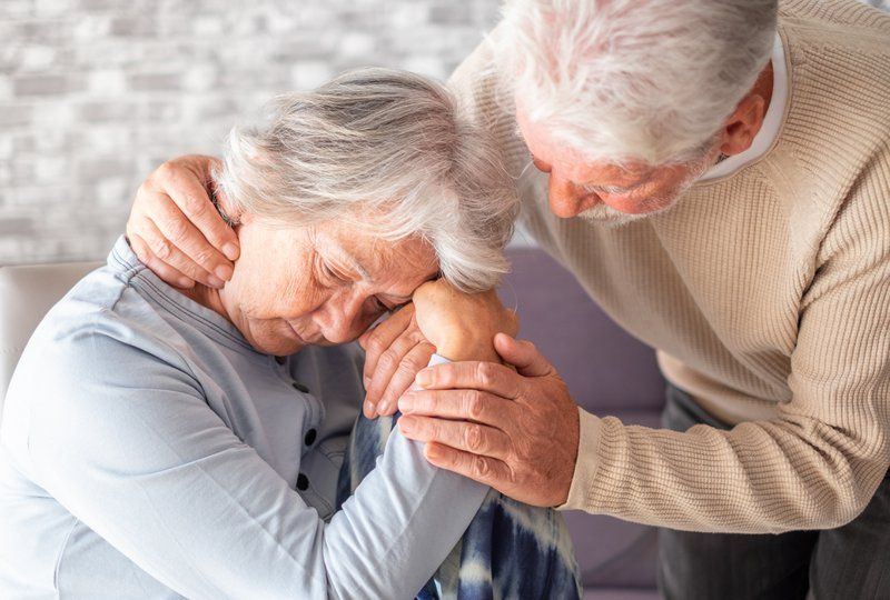 An elderly man comforts an elderly woman who appears distressed, sitting with her head resting on her arms while he gently embraces her, highlighting the importance of supporting seniors and promoting mental health awareness.