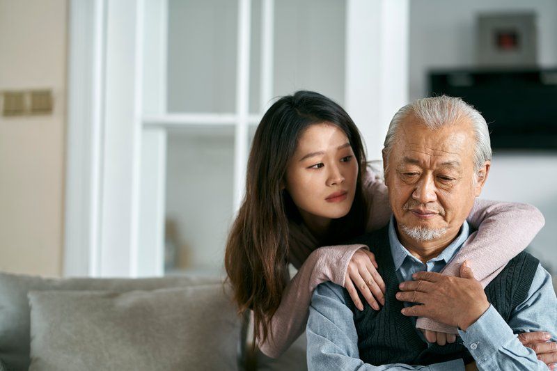 A young woman stands behind an older man, resting her arms on his shoulders as they sit on a couch. The man looks down thoughtfully while the woman gazes at him with concern, offering support as they cope with a recent Dementia diagnosis.