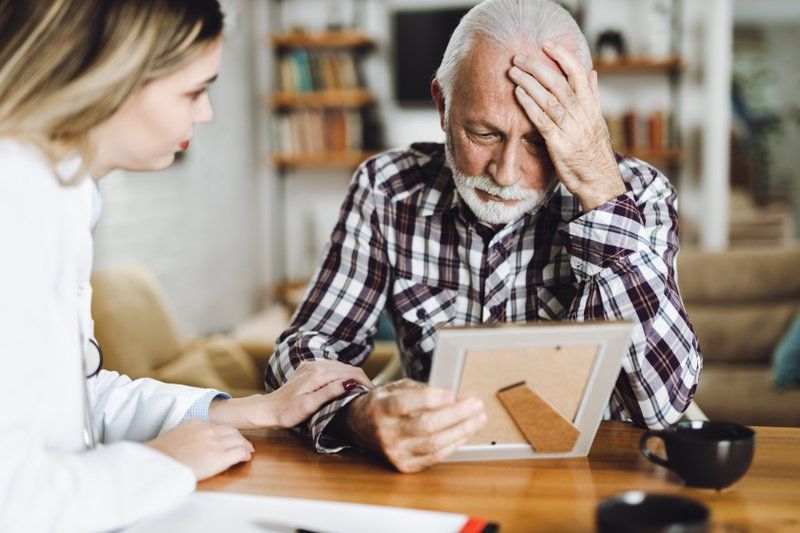 An elderly man holds a picture frame, looking distressed, while a woman beside him, possibly a caregiver or doctor, offers support—highlighting the importance of memory care in ensuring comfort and understanding for seniors.