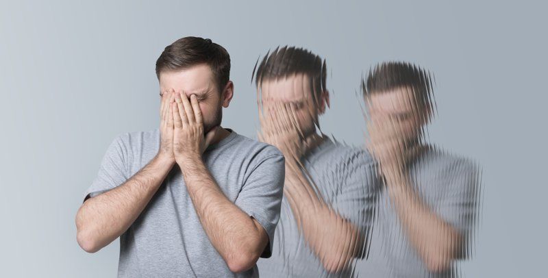 A man in a grey t-shirt covers his face with his hands; a blurred, duplicated effect suggests dizziness or confusion, often seen with Lewy Body Dementia and its associated hallucinations.