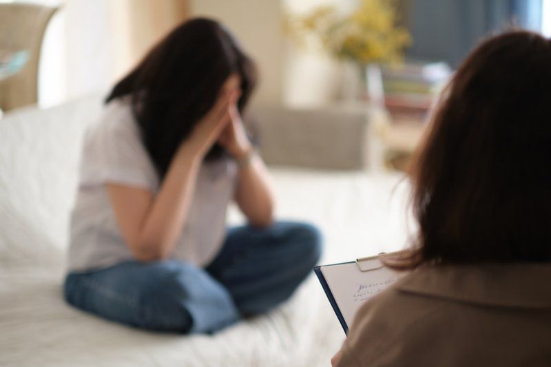 A person sits on a couch covering their face with their hands while another person with a clipboard faces them, suggesting a therapy or counseling session focused on Alzheimer's care.