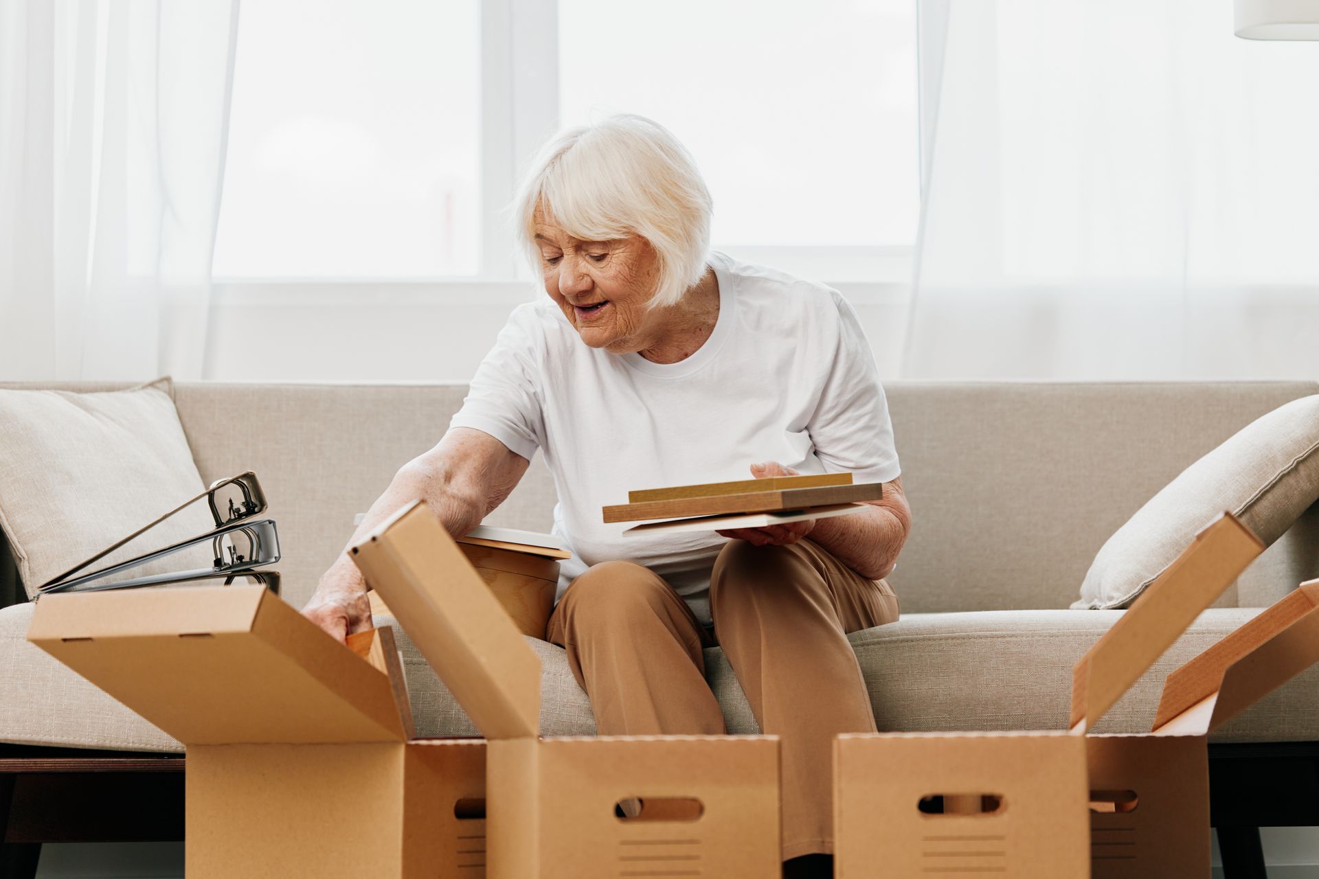 An elderly woman sits on a sofa, packing items into cardboard boxes in a well-lit room as she prepares for her move to assisted living.