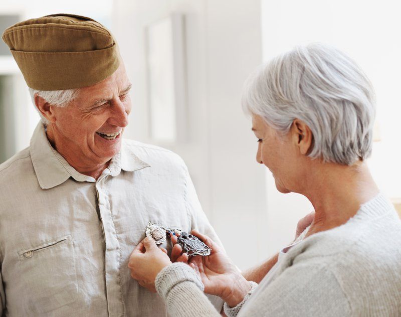 An older man in a military cap smiles as a woman pins medals onto his shirt inside a bright room, celebrating veterans and the bonds they share with their families at Assured Assisted Living.
